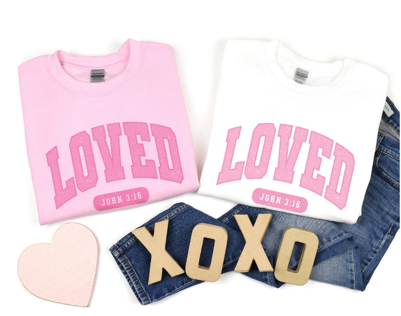 Op de afbeelding: Twee roze sweatshirts met het woord "LOVED" in roze letters en de tekst "JOHN 3:16" eronder. De sweatshirts liggen op een witte achtergrond met een roze hart en houten letters die "XOXO" spellen.