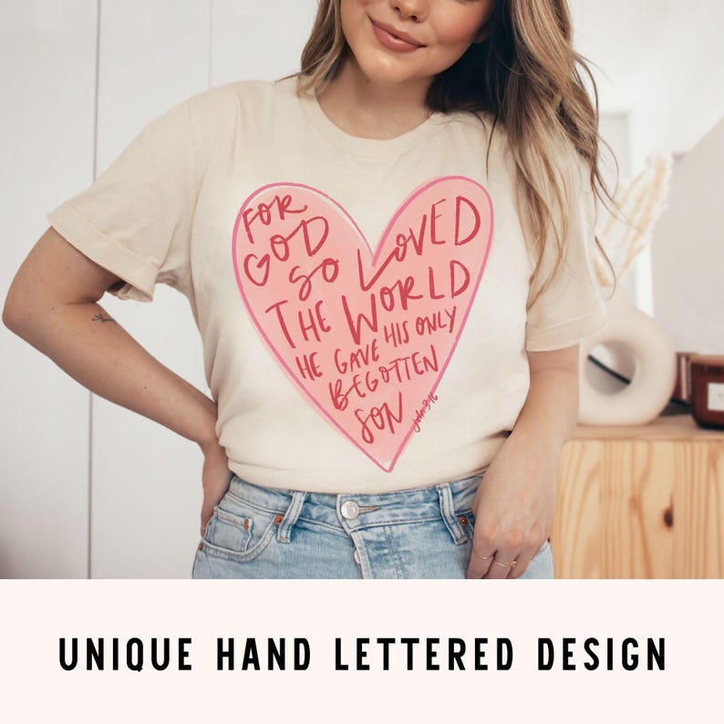 Puede incluir: Una camiseta beige con un gr&aacute;fico de coraz&oacute;n rosa. El coraz&oacute;n tiene el texto "For God so loved the world, he gave his only begotten son" en su interior. El texto est&aacute; escrito a mano.