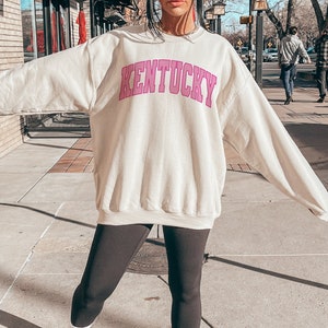 May include: A cream colored sweatshirt with the word "Kentucky" printed in pink on the front. The sweatshirt is worn by a person standing on a sidewalk.
