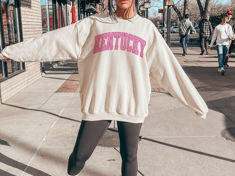 May include: A cream colored sweatshirt with the word "Kentucky" printed in pink on the front. The sweatshirt is worn by a person standing on a sidewalk.
