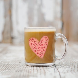 Puede incluir: Una taza de vidrio transparente con una pegatina en forma de coraz&oacute;n rosa que dice "Porque tanto am&oacute; Dios al mundo, que dio a su Hijo unig&eacute;nito". La taza est&aacute; llena de caf&eacute;.