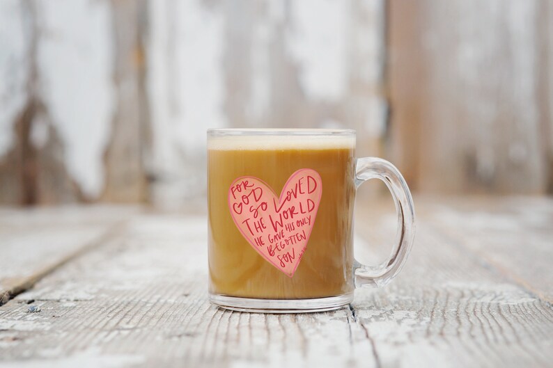 Puede incluir: Una taza de vidrio transparente con una pegatina en forma de coraz&oacute;n rosa que dice "Porque tanto am&oacute; Dios al mundo, que dio a su Hijo unig&eacute;nito". La taza est&aacute; llena de caf&eacute;.