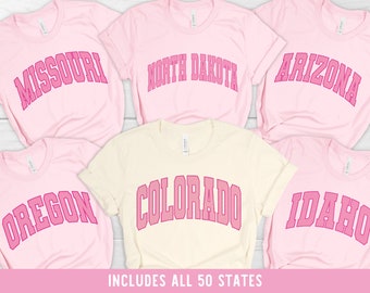 Op de afbeelding: Vijf roze T-shirts met verschillende staten in roze op de voorkant bedrukt. De staten zijn Missouri, North Dakota, Arizona, Oregon en Idaho. De tekst "INCLUDES ALL 50 STATES" staat onderaan de afbeelding.