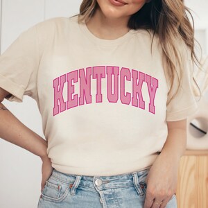 May include: A cream colored t-shirt with a pink arched graphic that says "KENTUCKY".