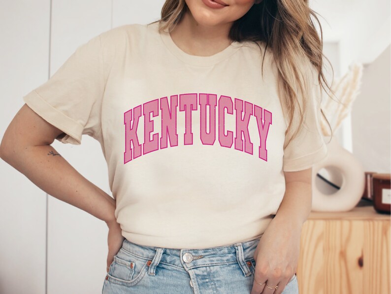May include: A cream colored t-shirt with a pink arched graphic that says "KENTUCKY".
