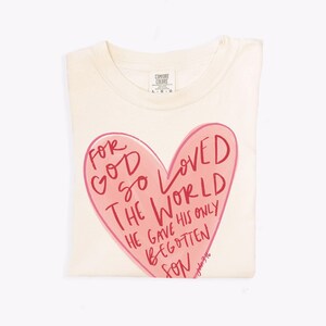 Puede incluir: Una camiseta de color crema con un gr&aacute;fico de coraz&oacute;n rosa. El coraz&oacute;n tiene las palabras "For God so loved the world" escritas en rojo. El texto contin&uacute;a debajo del coraz&oacute;n, "He gave his only begotten son." El texto "John 3:16" est&aacute; escrito en letras rojas m&aacute;s peque&ntilde;as debajo del coraz&oacute;n.