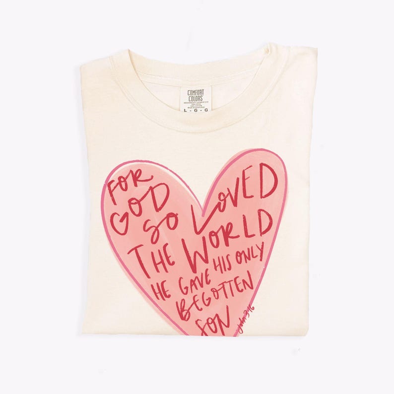 Puede incluir: Una camiseta de color crema con un gr&aacute;fico de coraz&oacute;n rosa. El coraz&oacute;n tiene las palabras "For God so loved the world" escritas en rojo. El texto contin&uacute;a debajo del coraz&oacute;n, "He gave his only begotten son." El texto "John 3:16" est&aacute; escrito en letras rojas m&aacute;s peque&ntilde;as debajo del coraz&oacute;n.