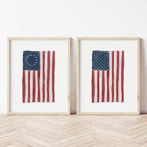 Puede incluir: Dos impresiones enmarcadas de la bandera estadounidense. Una presenta un círculo de estrellas sobre un fondo azul, la otra un campo de estrellas. Ambas tienen rayas rojas y blancas. Los marcos son de madera clara.