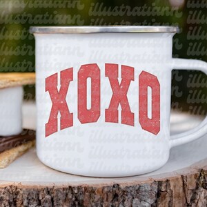 May include: White enamel camping mug with a red XOXO graphic. The mug is sitting on a wooden surface.
