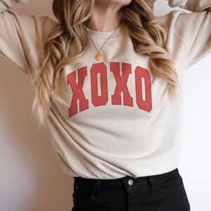 May include: A beige sweatshirt with a red "XOXO" graphic printed on the front. The sweatshirt has long sleeves and a crew neck.