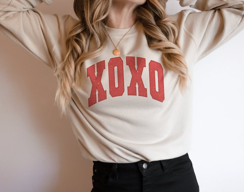 May include: A beige sweatshirt with a red "XOXO" graphic printed on the front. The sweatshirt has long sleeves and a crew neck.