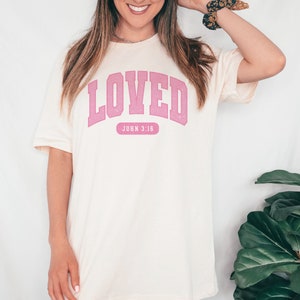 Op de afbeelding: Een wit T-shirt met een roze "LOVED"-afbeelding en de tekst "JOHN 3:16" eronder.
