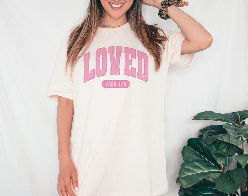Op de afbeelding: Een wit T-shirt met een roze "LOVED"-afbeelding en de tekst "JOHN 3:16" eronder.
