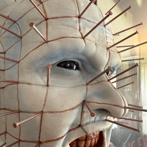 Może przedstawiać: Szczegółowe zbliżenie bladej, wyrzeźbionej twarzy z licznymi gwoździami w kolorze miedzi. Twarz ma wzór kratki i szeroko otwarte usta odsłaniające zęby. Oczy są ciemne, a ogólny efekt jest niepokojący.