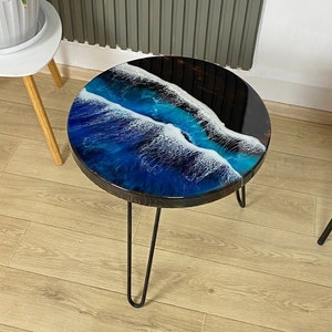 Może przedstawiać: Okrągły stolik boczny z czarną metalową ramą i nogami typu hairpin. Blat stołu zdobi niebiesko-biały wzór fal oceanu z czarnymi akcentami. Stół ma średnicę około 46 cm.