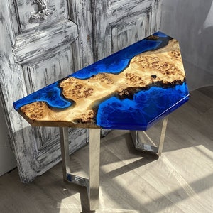 May include: A distinctive side table with a live edge wooden top incorporating a blue epoxy resin river design. The table has a natural wood grain pattern with a glossy finish and is supported by brushed metal legs. The table's dimensions are approximately 76 cm long, 46 cm wide, and 51 cm high.