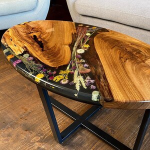 May include: Oval-shaped coffee table with a wooden top and black epoxy resin inlay featuring pressed flowers. The table has a black metal X-shaped base.
