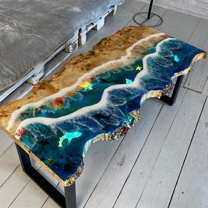 Pode incluir: Uma mesa de centro única com um design de praia. A mesa apresenta um tampo de madeira com uma cena oceânica de resina, completa com ondas, vida marinha e uma praia arenosa. A mesa é suportada por pernas de metal preto.