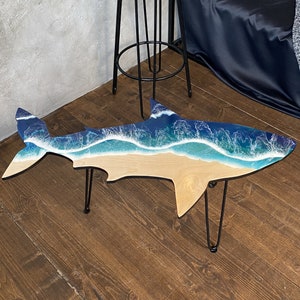May include: A wooden shark-shaped coffee table with a blue and white resin ocean wave design. The table has black hairpin legs and is sitting on a wooden floor.