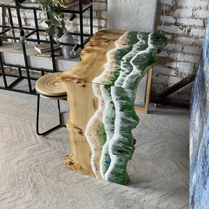 May include: A unique live edge table with a beach-themed epoxy resin design. The table features a natural wood grain on one side and a vibrant green and white resin wave pattern on the other. The table is supported by a natural wood base.