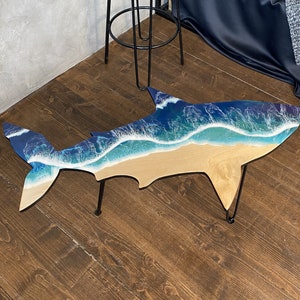 May include: A unique coffee table shaped like a shark with a blue and white resin ocean wave design. The table has black metal legs.