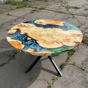 Puede incluir: Mesa redonda de madera con una incrustación de resina epoxi azul y verde. La mesa tiene una base en forma de X de metal negro.