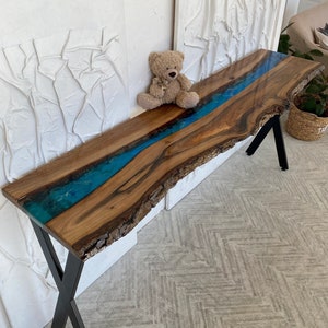 May include: A long console table featuring a natural live edge wood slab with a central river of translucent blue epoxy resin. The table is supported by black metal X-shaped legs and displays a prominent wood grain with dark brown tones.