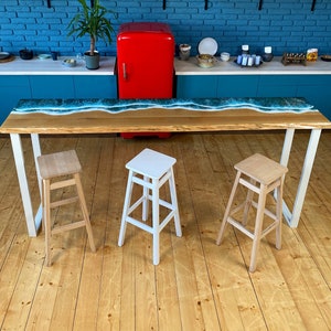 May include: A long wooden table with a blue epoxy resin wave design. The table has white metal legs and is set with three wooden stools. Two of the stools are natural wood and one is white.