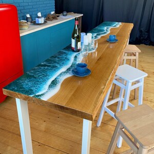 May include: A wooden table with a blue and white epoxy resin ocean wave design. The table has white metal legs and is set up with three wooden stools.