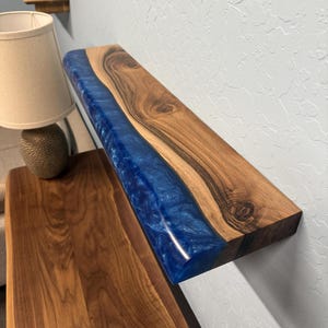 May include: A wooden shelf with a blue resin river running through the center. The wood has a natural grain pattern and is stained a warm brown. The blue resin has a glossy finish, creating a striking contrast.