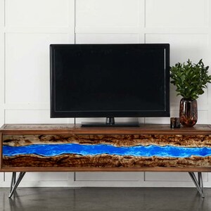 Puede incluir: Un mueble de televisión rectangular de madera con un diseño de borde natural y una incrustación de resina azul vibrante que recorre horizontalmente el centro. La unidad se apoya sobre cuatro patas metálicas inclinadas y sostiene una pantalla de televisión negra.
