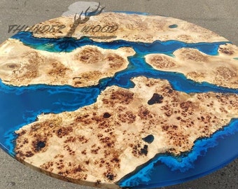May include: A round table top featuring a natural light-coloured wood burl with a complex, textured grain pattern integrated with a vibrant, translucent blue epoxy resin river design. The text THUNDER WOOD is visible on the surface of the wood.