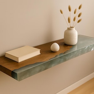 Puede incluir: Una estantería flotante moderna con una superficie de madera y un borde de resina transparente. Un libro cerrado, un jarrón blanco con flores secas y un objeto blanco en forma de huevo se exhiben en la estantería. La estantería está montada en una pared beige.