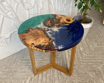 Op de afbeelding: Een ronde bijzettafel met een houten schijf met natuurlijke rand, geïntegreerd in teal en diepblauwe epoxyhars. Het oppervlak vertoont witte wervelingen in de hars en rust op drie lichtgekleurde houten poten die aan de basis zijn verbonden.