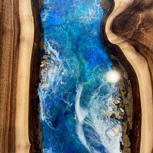 May include: A close-up of a wooden table with a blue and white epoxy resin inlay. The resin has a swirling pattern that resembles waves. The wood is a dark brown color with lighter brown streaks.