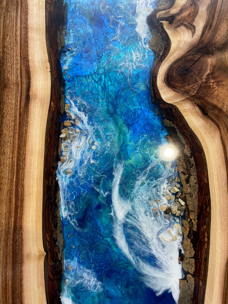 May include: A close-up of a wooden table with a blue and white epoxy resin inlay. The resin has a swirling pattern that resembles waves. The wood is a dark brown color with lighter brown streaks.