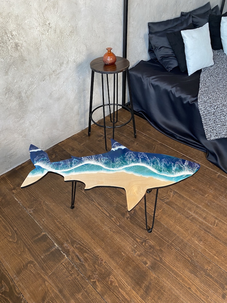 May include: A wooden shark-shaped coffee table with a blue and white resin ocean wave design. The table has black hairpin legs and is sitting on a wooden floor.