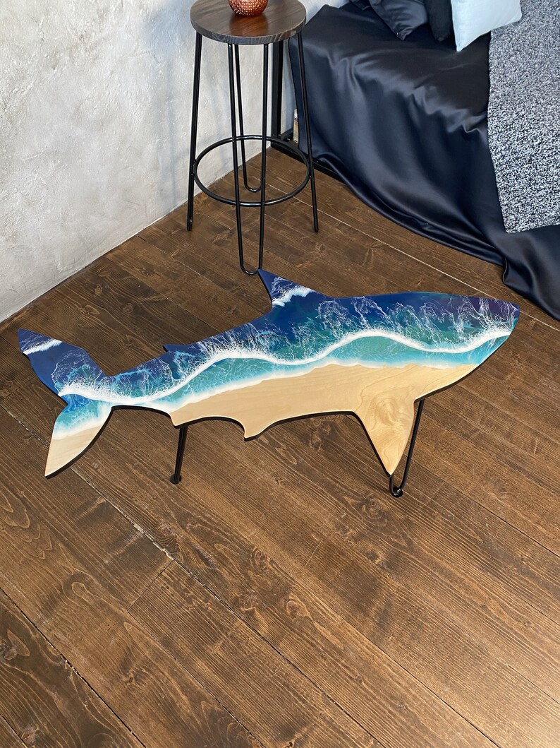 May include: A unique coffee table shaped like a shark with a blue and white resin ocean wave design. The table has black metal legs.