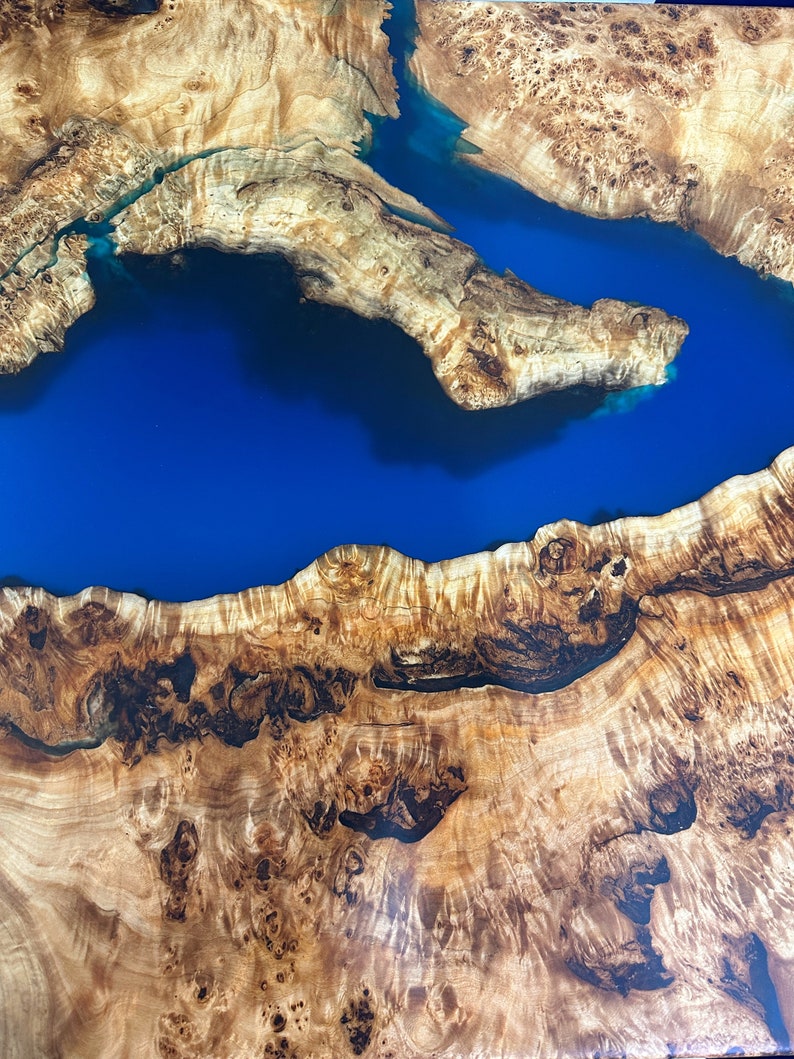 May include: A close-up view of a wooden table with a river of blue resin. The wood has a natural, light brown color with intricate patterns and knots. The blue resin creates a striking contrast, resembling a flowing river or lake.