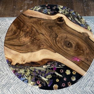 K&ouml;nnte beinhalten: Runder Couchtisch aus Holz mit einer Baumkante. Der Tisch hat eine dunkle Holzmaserung in der Mitte und einen Rand aus getrockneten Blumen, die in klarem Harz eingeschlossen sind. Auf einem hellgrauen Teppich.