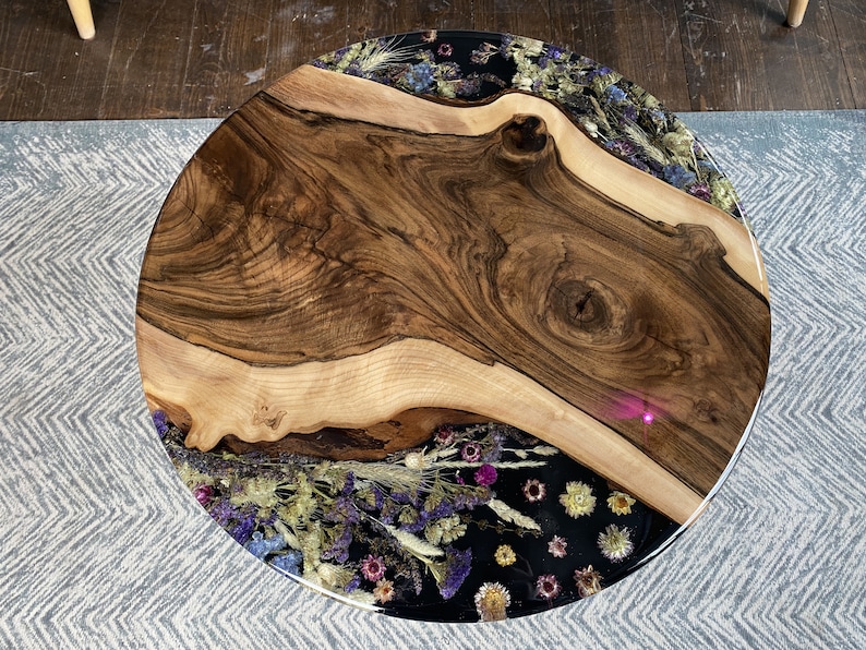 K&ouml;nnte beinhalten: Runder Couchtisch aus Holz mit einer Baumkante. Der Tisch hat eine dunkle Holzmaserung in der Mitte und einen Rand aus getrockneten Blumen, die in klarem Harz eingeschlossen sind. Auf einem hellgrauen Teppich.