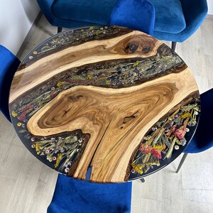 May include: Round wooden table with a clear resin top inlaid with dried flowers. The table has a natural wood grain pattern and is surrounded by blue chairs and a blue sofa.