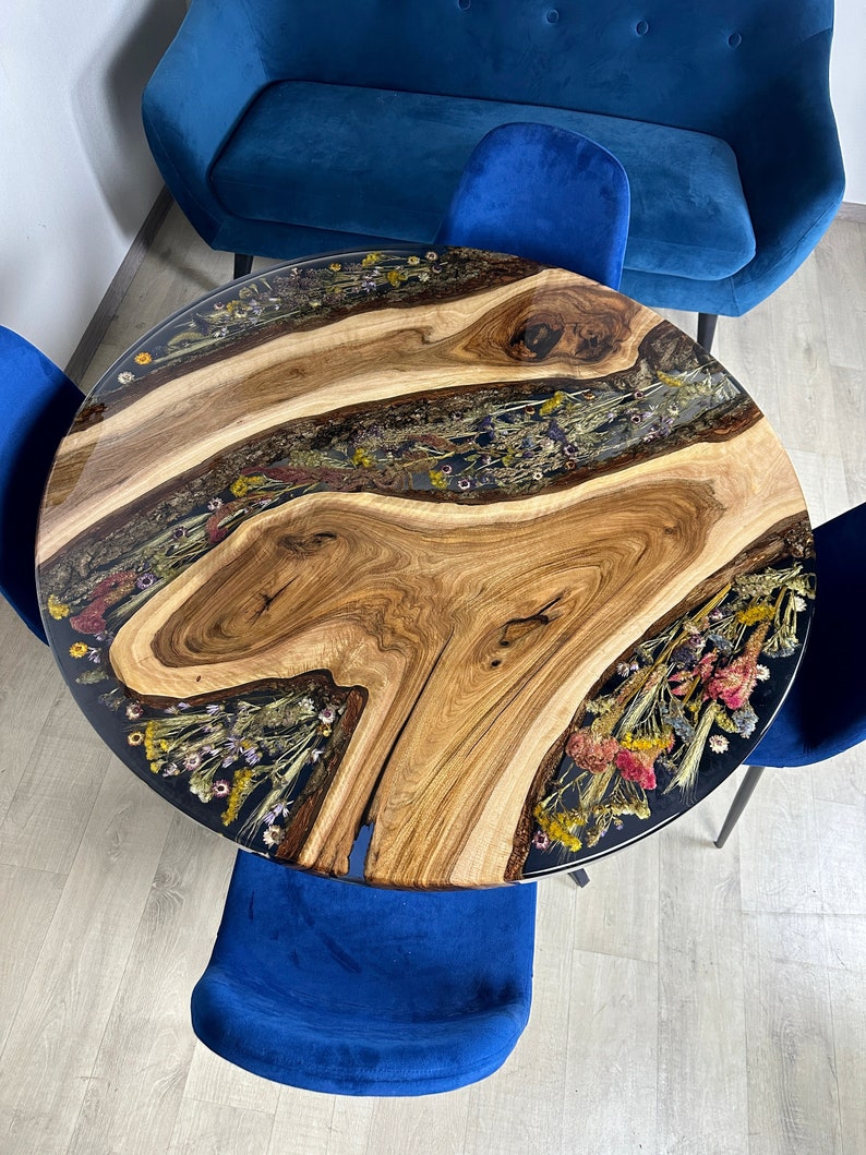 May include: Round wooden table with a clear resin top inlaid with dried flowers. The table has a natural wood grain pattern and is surrounded by blue chairs and a blue sofa.