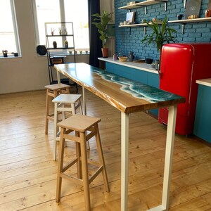 May include: A wooden bar table with a blue and white epoxy resin top, featuring a wave design. The table has white metal legs and is set against a blue brick wall. Three wooden bar stools are placed near the table.