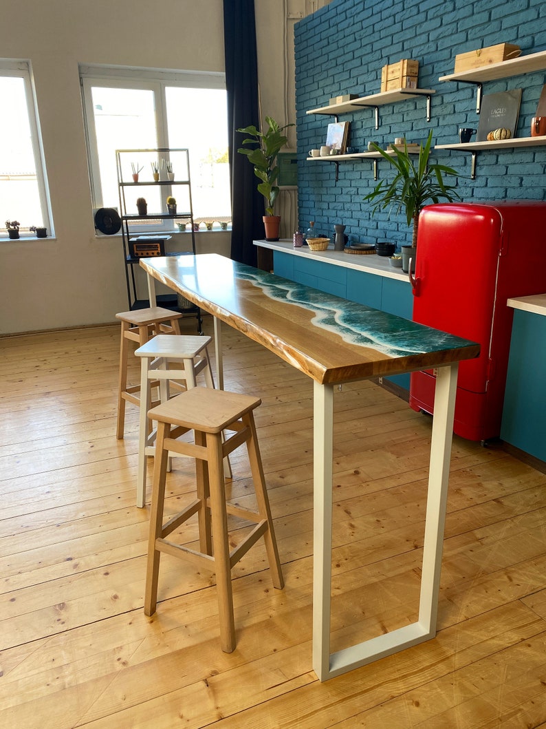 May include: A wooden bar table with a blue and white epoxy resin top, featuring a wave design. The table has white metal legs and is set against a blue brick wall. Three wooden bar stools are placed near the table.
