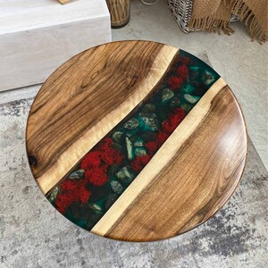 Puede incluir: Una mesa redonda con dos tablones de madera natural con vetas marcadas, separados por una incrustaci&oacute;n central tipo r&iacute;o. La incrustaci&oacute;n contiene resina transl&uacute;cida color verde azulado con piedras de r&iacute;o grises y musgo rojo brillante.