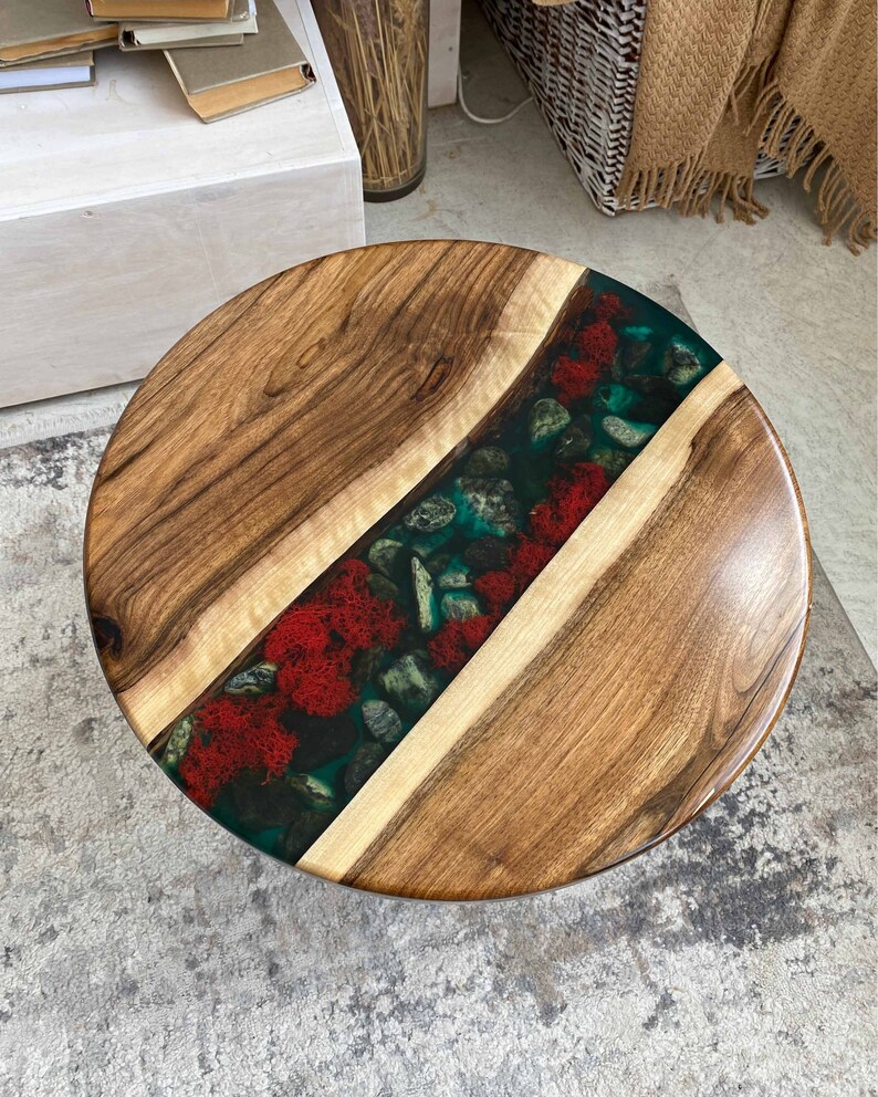 Puede incluir: Una mesa redonda con dos tablones de madera natural con vetas marcadas, separados por una incrustaci&oacute;n central tipo r&iacute;o. La incrustaci&oacute;n contiene resina transl&uacute;cida color verde azulado con piedras de r&iacute;o grises y musgo rojo brillante.
