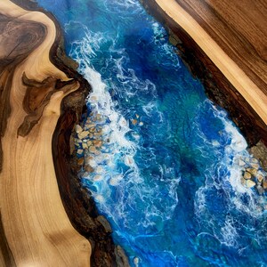May include: A close-up of a wooden table top with a blue and white epoxy river running through the center. The epoxy has a swirling pattern that resembles waves and includes white accents that look like foam.