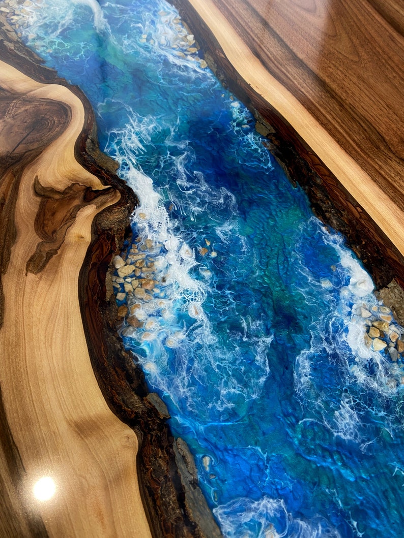 May include: A close-up of a wooden table top with a blue and white epoxy river running through the center. The epoxy has a swirling pattern that resembles waves and includes white accents that look like foam.
