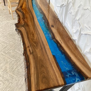 May include: A long console table featuring a live edge walnut wood slab with a central river design made of translucent blue epoxy resin. The surface is polished to a high gloss, showcasing the natural wood grain and dark brown tones.